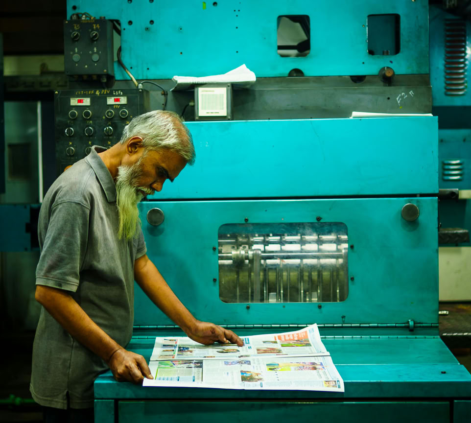 Un homme &acirc;g&eacute; examine des documents fra&icirc;chement imprim&eacute;s devant une grande presse &agrave; imprimer turquoise.